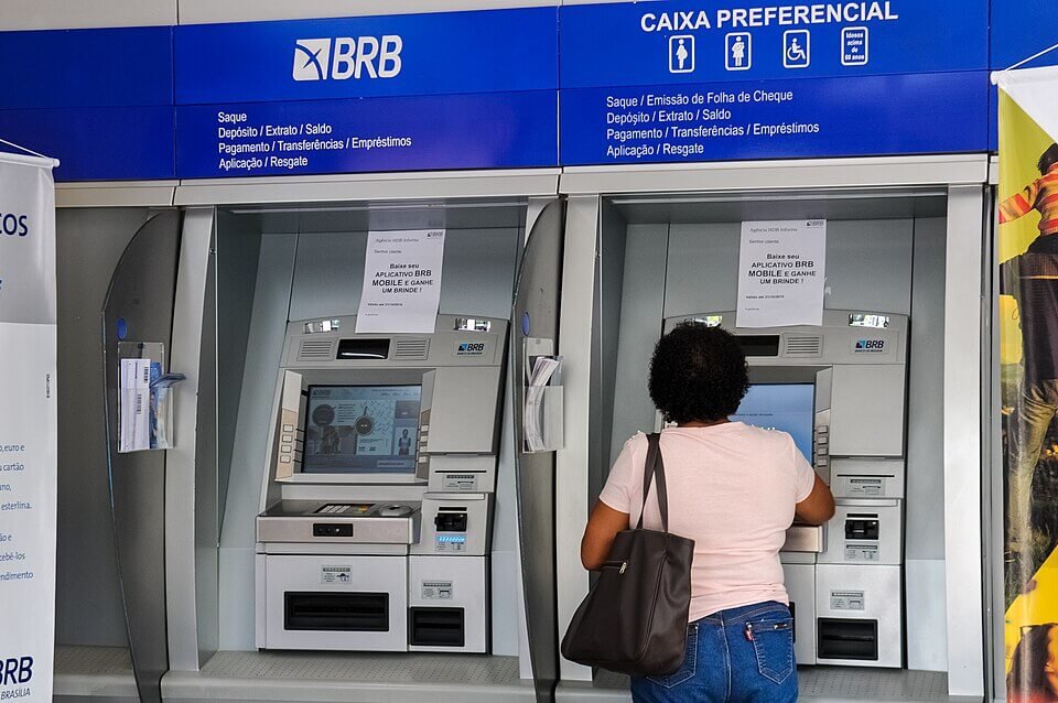 Duas máquinas de caixas eletrônicos integradas na parede azul dentro de uma agência do BRB (Banco de Brasília), com uma pessoa de costas utilizando um dos terminais de atendimento automático.